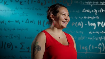 A woman in a red sleeveless top stands in from of a wall of mathematical equations. Photo credit: Jordan Stead / Amazon.