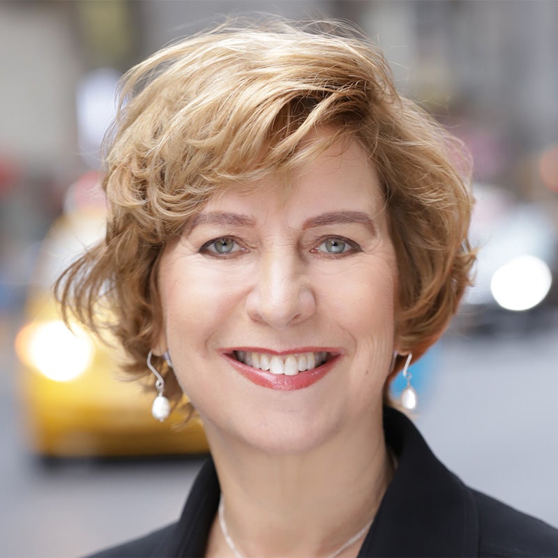 Vivian Shchiller's headshot. A person with short hair wearing long earrings stands beside a road smiling at the camera. A taxi is visible over their shoulder.