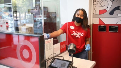Photo: A smiling Target team member wearing a red Target shirt, black face mask and blue gloves, standing behind a Plexiglass shield at the Order Pickup counter