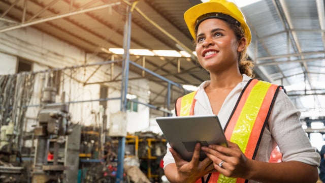 Photo of a young woman factory worker wearing a hard hat and reflector vest. She is smiling and holding an electronic tablet.