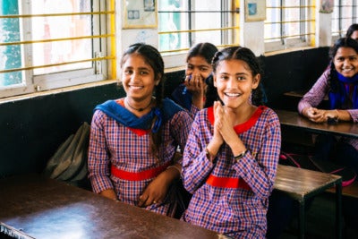 A group of young students sit at wooden desks smiling at the camera.