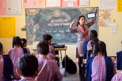 A group of students sit at individual desks facing the front of the classroom. A teacher stands in front of a chalk board with vibrant drawings that reads, "Girl Rising." The teacher holds up a computer tablet and talks to the class.