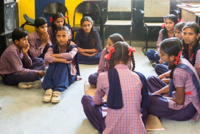 A group of students sit on the floor of a classroom chatting.