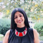 Nidhi Shukla's headshot. A person with long hair wearing a vibrant necklace stands smiling at the camera. Trees and shrubs are in the background.