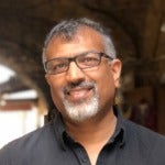 Shahed Amanullah's headshot. A person with a goatee and wearing glasses stands under an archway smiling at the camera.
