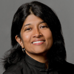 Sampriti Ganguli's headshot. A person with medium-length hair that's tucked behind one ear and is wearing a high-necked shirt stands in front of a wall smiling at the camera.