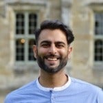 Adam Motiwala's headshot. Image depicts a person with short hair, a beard, and a mustache wearing a shirt with buttons smiling at the camera.