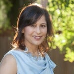 Anju Sharma's headshot. Image depicts a person with shoulder-length hair wearing a sleeveless blouse outside smiling at the camera.