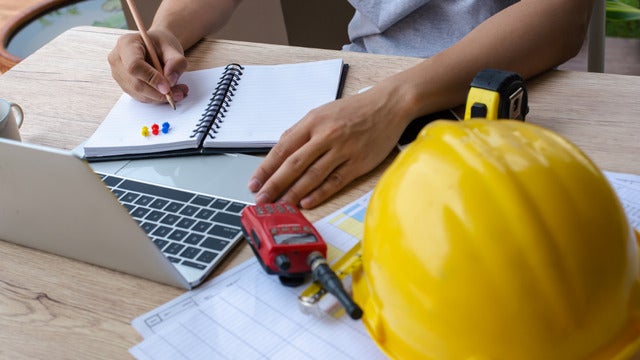 A photo of a man working at a desk with a hard hat, a laptop, and a handheld radio in front of him