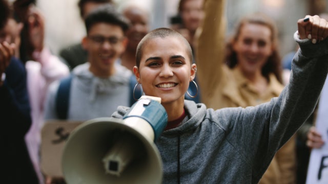Photo of a young woman leading a protest. She has one fist in the air, while holding a megaphone in her other hand.