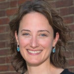 Elizabeth Hansen Shapiro's headshot. A person with shoulder-length hair wearing turquoise stone earrings is positioned in front of a brick wall smiling at the camera.