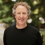 Jim Friedlich's headshot. A person with short hair wearing a t-shirt is outside, trees in the background, smiling at the camera.