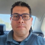 Joey Gardea's headshot. A person with short hair wearing glasses and a polo sits in a car wearing their seatbelt looking at the camera.