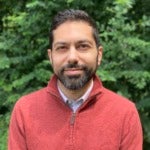 Kasem Rodriquez Mohsen's headshot. Image depicts a person with short hair, a beard, and a mustache wearing a quarter-zip sweater positioned in front of a shrub smiling at the camera.