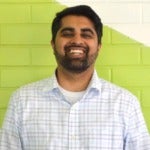 Khuram Zaman's headshot. Image depicts a person with short hair, a beard, and a mustache wearing a collared shirt positioned in front of a colorful brick wall smiling at the camera.