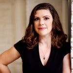 Laura Walker-McDonald's headshot. Image depicts a person with medium-length wavy hair wearing a v-neck t-shirt and necklace leaning against a brick wall, hand on their hip, looking at the camera.