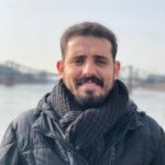 Oussama Abdellah Benhmida's headshot. Image depicts a person with short hair, a beard, and a mustache wearing a coat and scarf outside smiling at the camera.