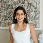 Prenra Mukharya's headshot. Image depicts a person with long hair wearing a tank top and earrings leaning against a table, a painting in the background, smiling at the camera.
