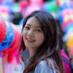 Xing Liu's headshot. A person with long hair wearing a collared shirt surrounded by colorful lanterns turns and smiles at the camera.