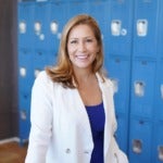 White woman with blonde hair stands in a white blazer smiling and looking straight at the camera