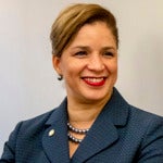 Smiling Black woman in a blue blazer stands with her arm crossed looking to the side of the camera.