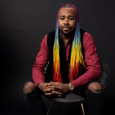 Jarvis Sam's headshot. A person with long, rainbow hair and a goatee wearing a collared shirt, vest, and ripped jeans sits in a chair with their hands clasped together, looking at the camera.