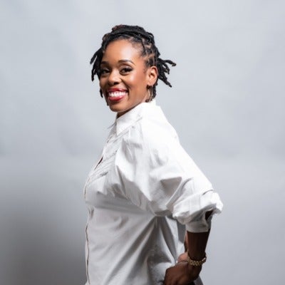Shijuade Kadree's headshot. A person with their hair pulled back who is wearing earrings and a collared shirt stands sideways with their hands on their hips, smiling at the camera.