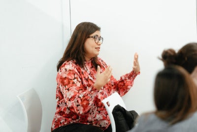 A woman with shoulder-length straight brown hair, glasses, and a pink floral long sleeve shirt sits against a white backdrop. She is speaking and gesturing with her hands.