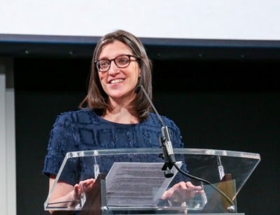 A white woman with shoulder length straight brown hair, glasses, and a navy shirt stands at a glass podium. She is smiling and looking to her left into the crowd. 