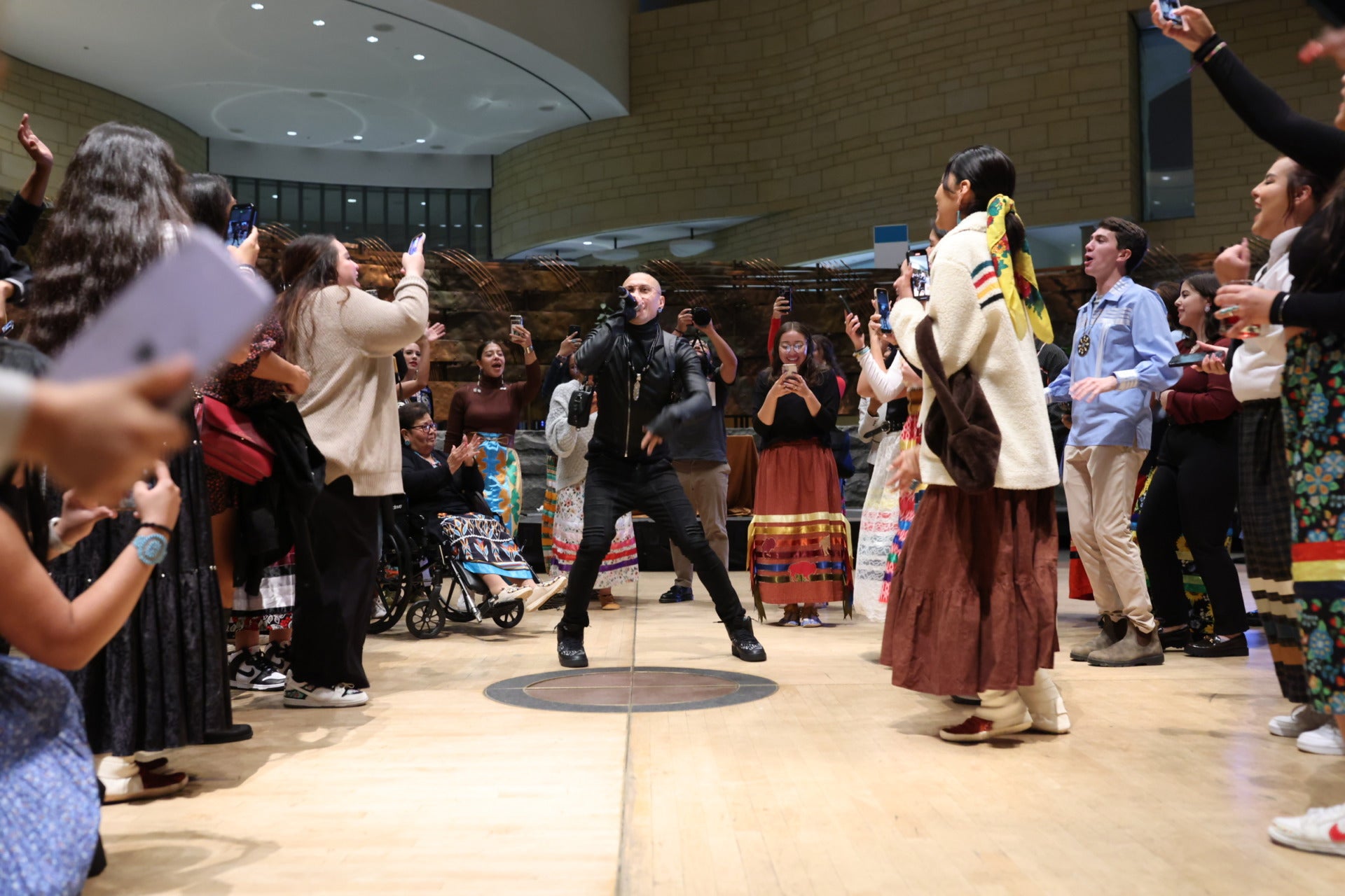 A crowd of Indigenous people wearing traditional attire gather around a singer who is jumping up in the air and holding a mic. He is wearing all black. The people surrounding him are also jumping, smiling, clapping, and being joyful. They are on a light brown floor.