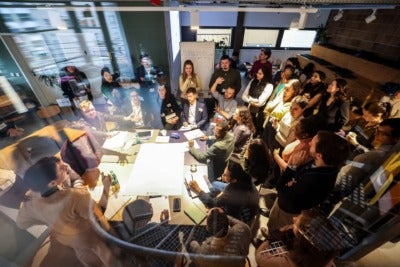 A large group of people standing around a table with a light shining down on it. They are excitedly talking, collaborating, and writing. The photo is taken on a stairwell above the table, looking down on everyone.