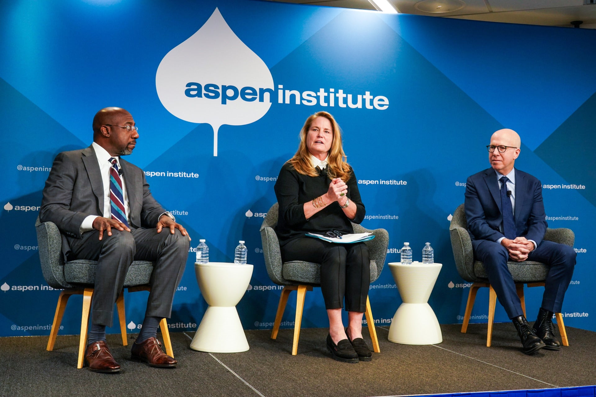 Three panelists are seated on a stage with white tables between them and water bottles on the tables. They are in front of a blue background with the Aspen Institute logo. On the left is Senator Ralph Warnock, a black man wearing a gray suit, white shirt, and colorful tie. In the middle is the Aspen Institute's Anne Mosle, a white woman with long blonde hair wearing a sweater and white collared shirt. To her right is the final panelist, author Jonathan Eig - a bald white man with glasses who is wearing a dark blue suit and tie.
