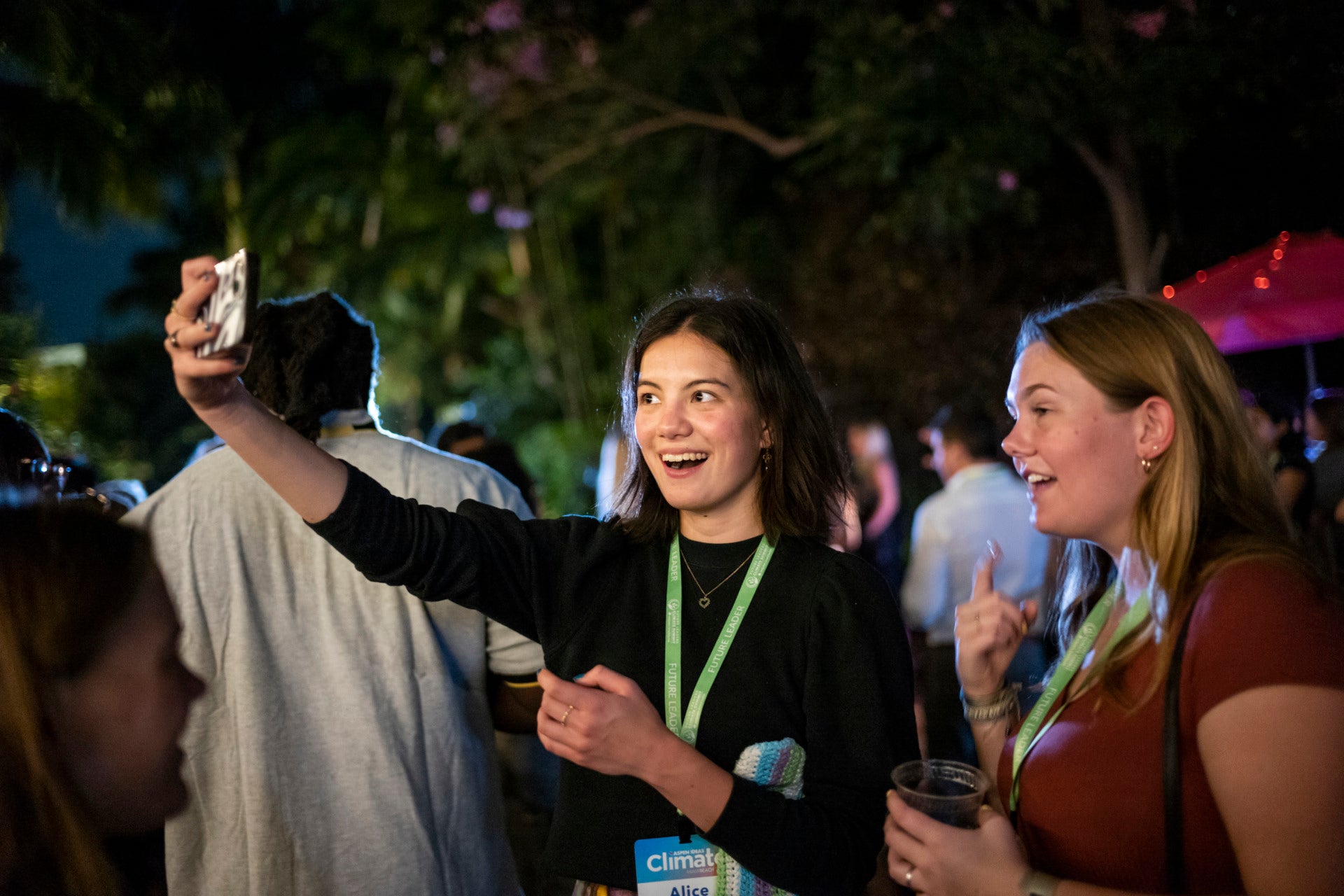 Two young women take a selfie together. They are amidst a large crowd outside at night time, and behind them are tall trees.