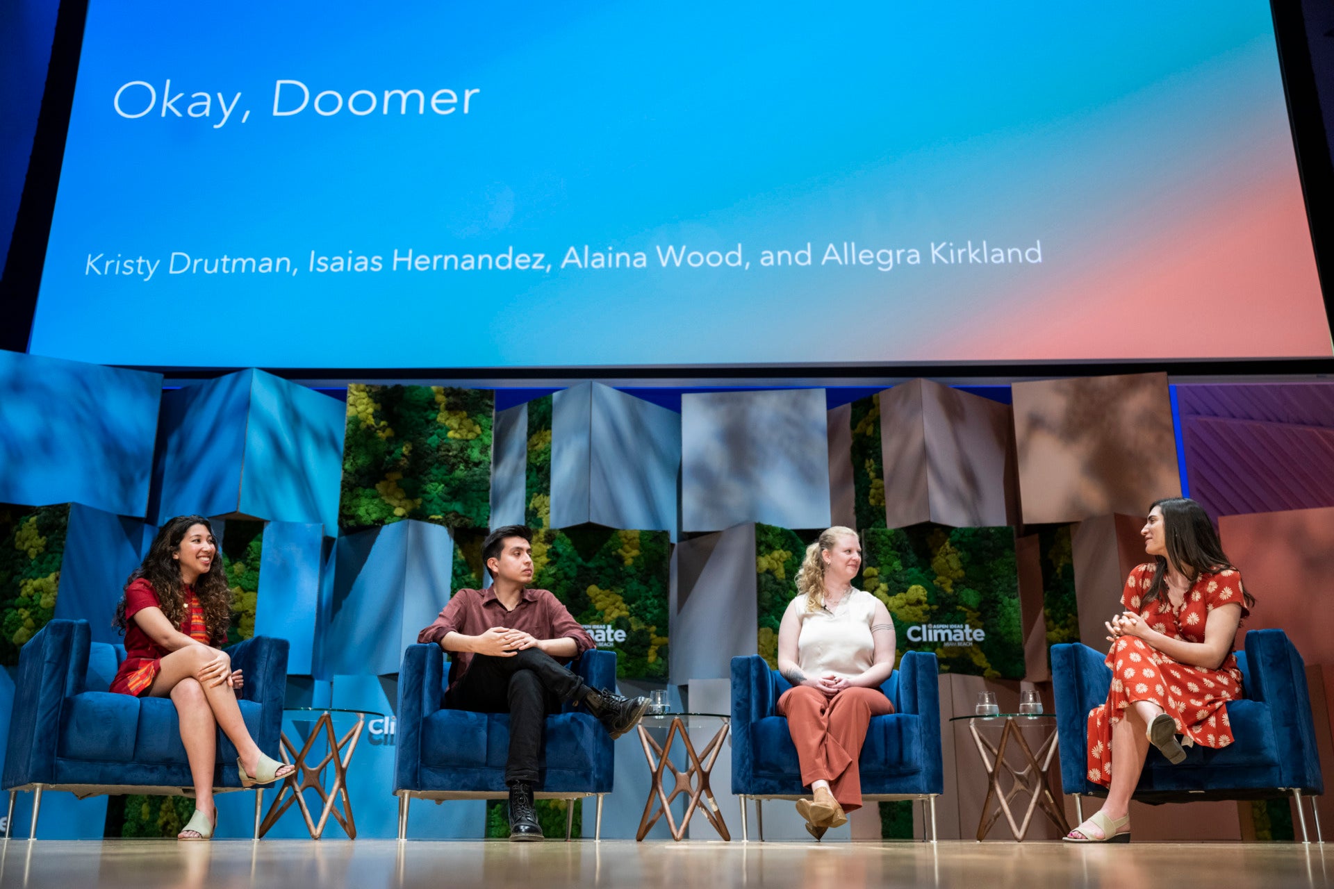 Four people sit in blue velvet armchairs on a colorful stage. Above them, a slide is projected that reads: "Okay, Doomer," followed by the names of the panelists. The panelists are looking to their left, listening intently to a fellow speaker.
