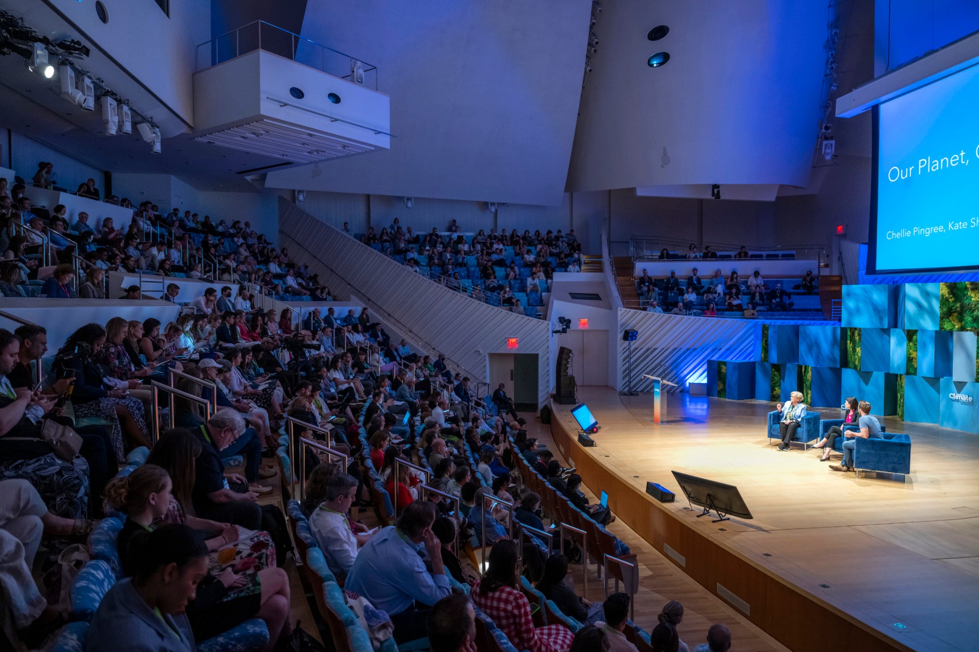 A large crowd sits in stadium style seating in an auditorium, looking at a stage where three people sit in blue velvet chairs having a discussion.