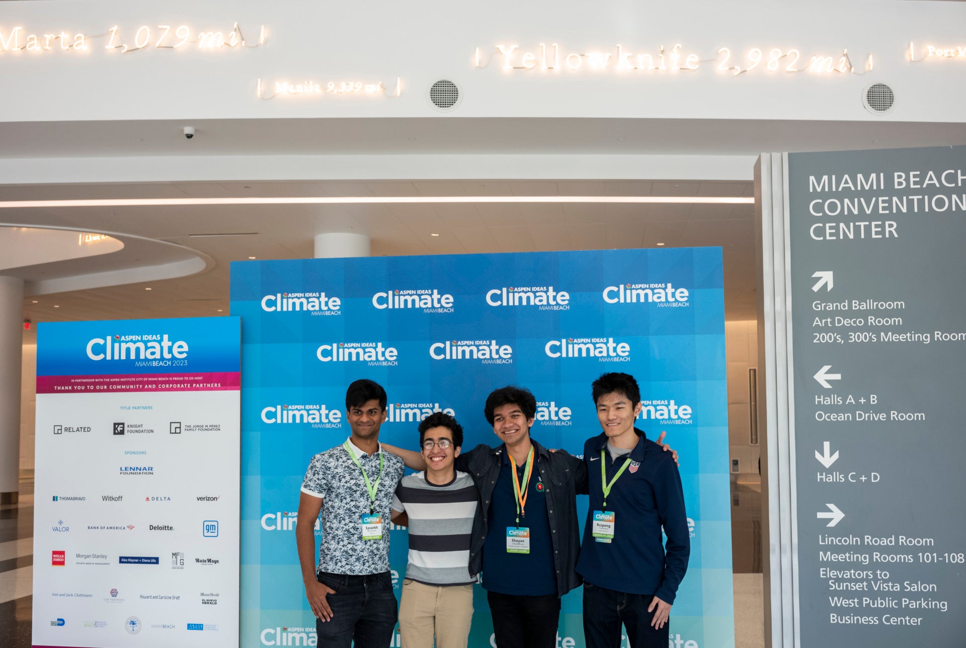 Four young adults stand in front of a step and repeat with a blue background and the Aspen Ideas Climate logo. To their left is a directional sign for navigating the Miami Beach Convention Center.