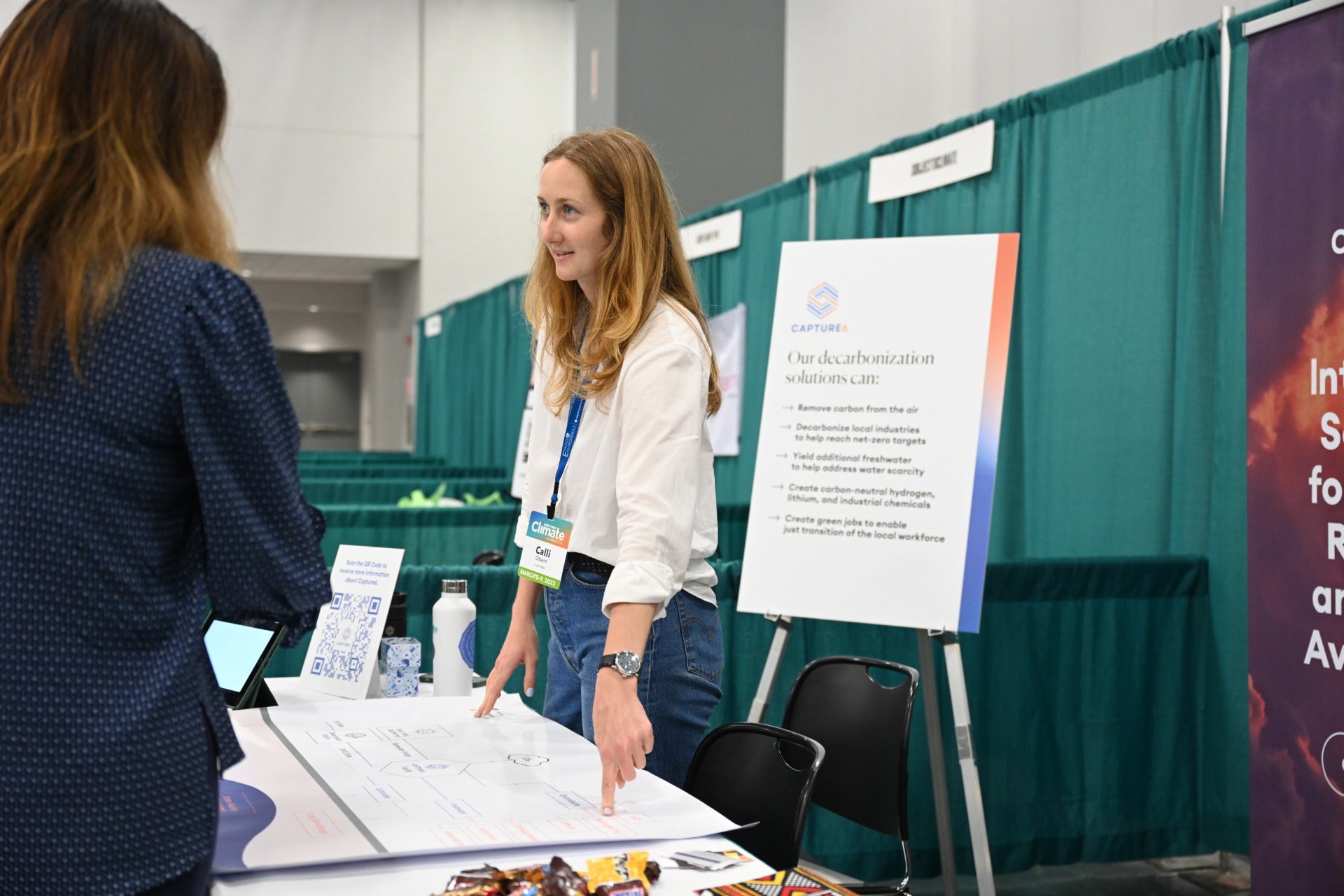 A woman with blonde hair stands behind a table and points to a drawing on the table while looking at a person standing in front of her.