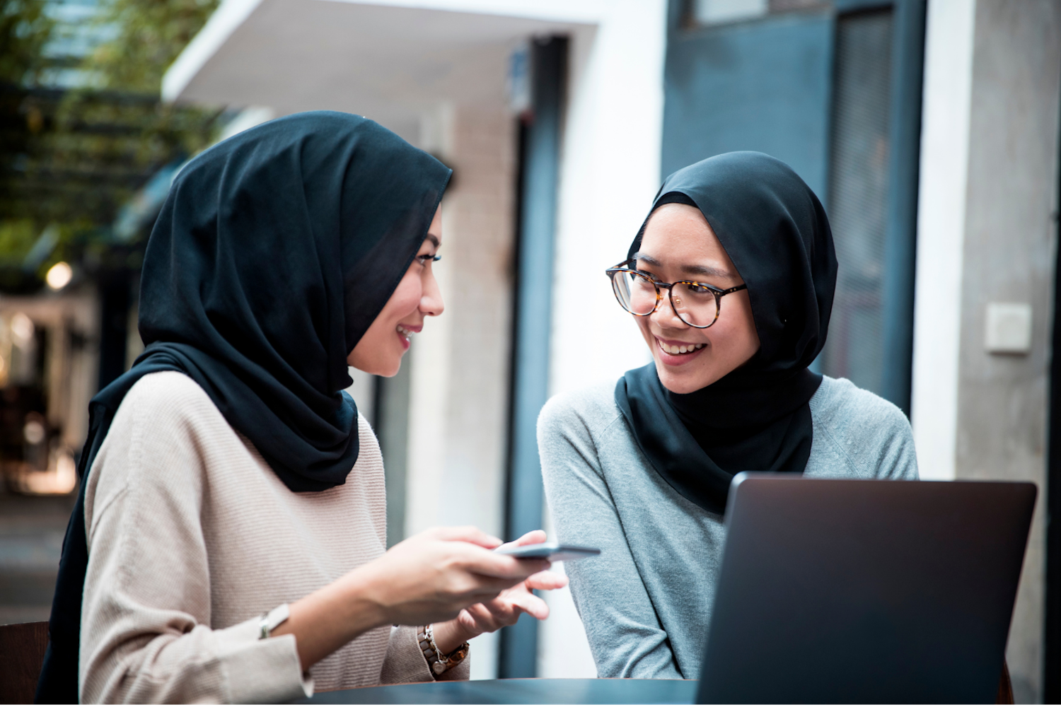 A photo of two Malaysians in head coverings smile and look at each other while they look at a laptop computer. One holds a phone.