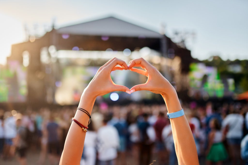 Concertgoer forms heart shape with hands at beachside music festival, crowd enjoys live performance in sunset, love symbol gesture at summer event, youth celebration of art outdoor entertainment.