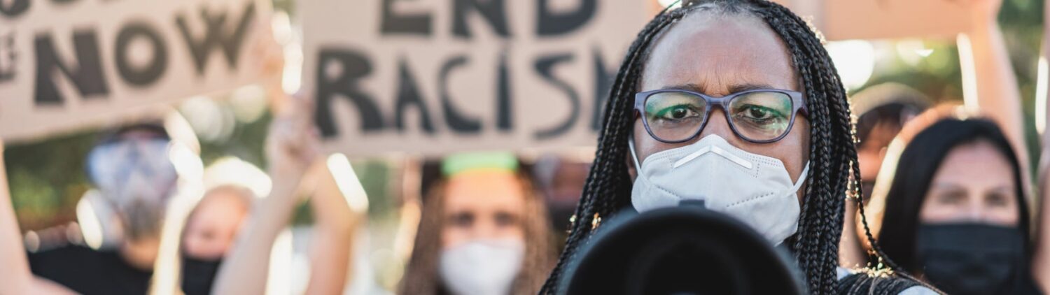 Photo of protestors holding signs like "Justice Now" and "End Racism." A Black woman wearing glasses and a face mask speaks through a megaphone at the front of the group.