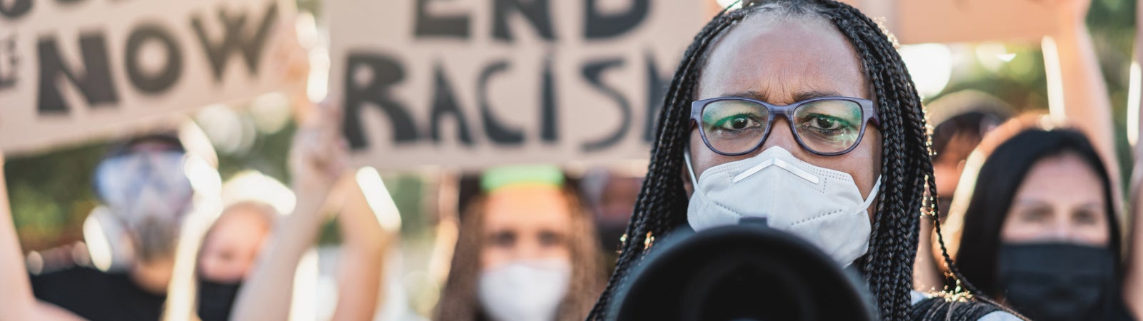 Photo of protestors holding signs like "Justice Now" and "End Racism." A Black woman wearing glasses and a face mask speaks through a megaphone at the front of the group.