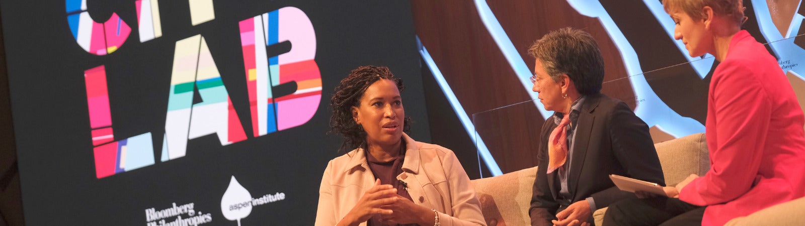 Three women sit on a stage for a panel, with a large colorful sign that says "City Lab" and the Bloomberg and Aspen Institute logos behind them. The woman on the far left, DC Mayor Muriel Bowser, is black with short hair and is wearing a tan button down. She is speaking and gesturing with her hands. To her right are two other women. The middle one has short salt and pepper hair, glasses, a black blazer and a neckerchief on. To the far right is a woman with short blonde hair and a pink jacket. Both are listening to Mayor Bowser speak.