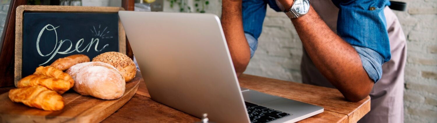 Photo of a Black bakery owner using a laptop. There is an "Open" sign on the counter, next to bread and pastries.