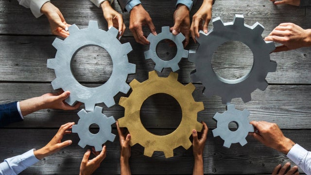 Photo of gears on a table, held together by the hands of people seated around it.