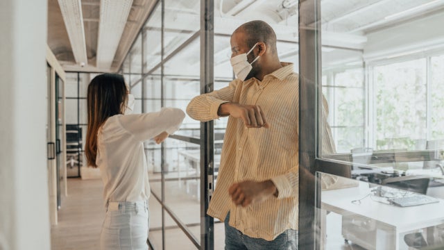 Photo of a Black man and Asian woman wearing face masks and bumping elbows in an office.