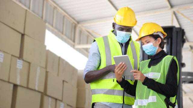 Photo of two warehouse workers wearing hard hats, reflector vests, and face masks. They are standing in front of a stack of cardboard boxes while looking at an electronic tablet. One is a Black man and the other is an Asian woman.