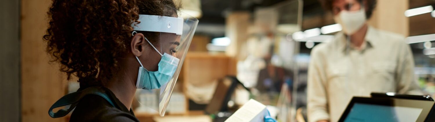 Photo of a young woman of color working at the checkout lane in a grocery store, wearing personal protective equipment, including a face mask, face shield, and gloves