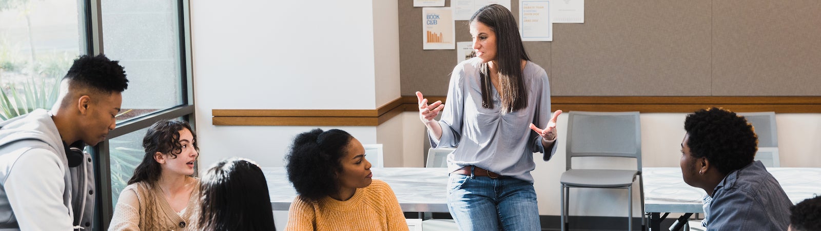 A photograph of a teacher leaning on a desk speaking to a group of high school students sitting around a table.