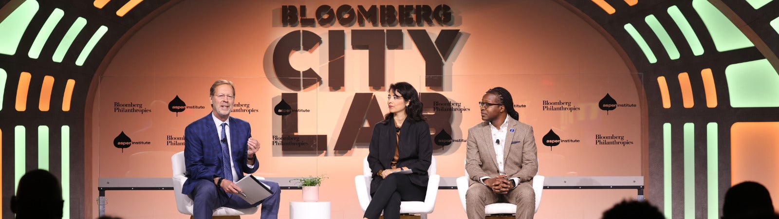 Three panelists sit on a stage with lights on each side and the words "Bloomberg CityLab" in large text behind them. On the left is Aspen Institute CEO Dan Porterfield, who is moderating. He speaks as the two panelists, a Hispanic woman with dark brown hair and a black pantsuit, and a Black man with his hair pulled back, glasses, and a tan suit sit with their hands in their laps listening.