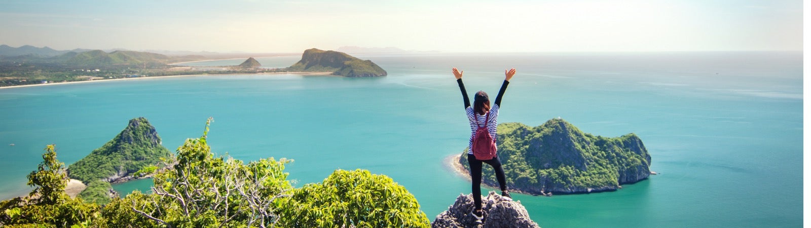 A woman standing on top of a mountain overlooking water with her arms raised.